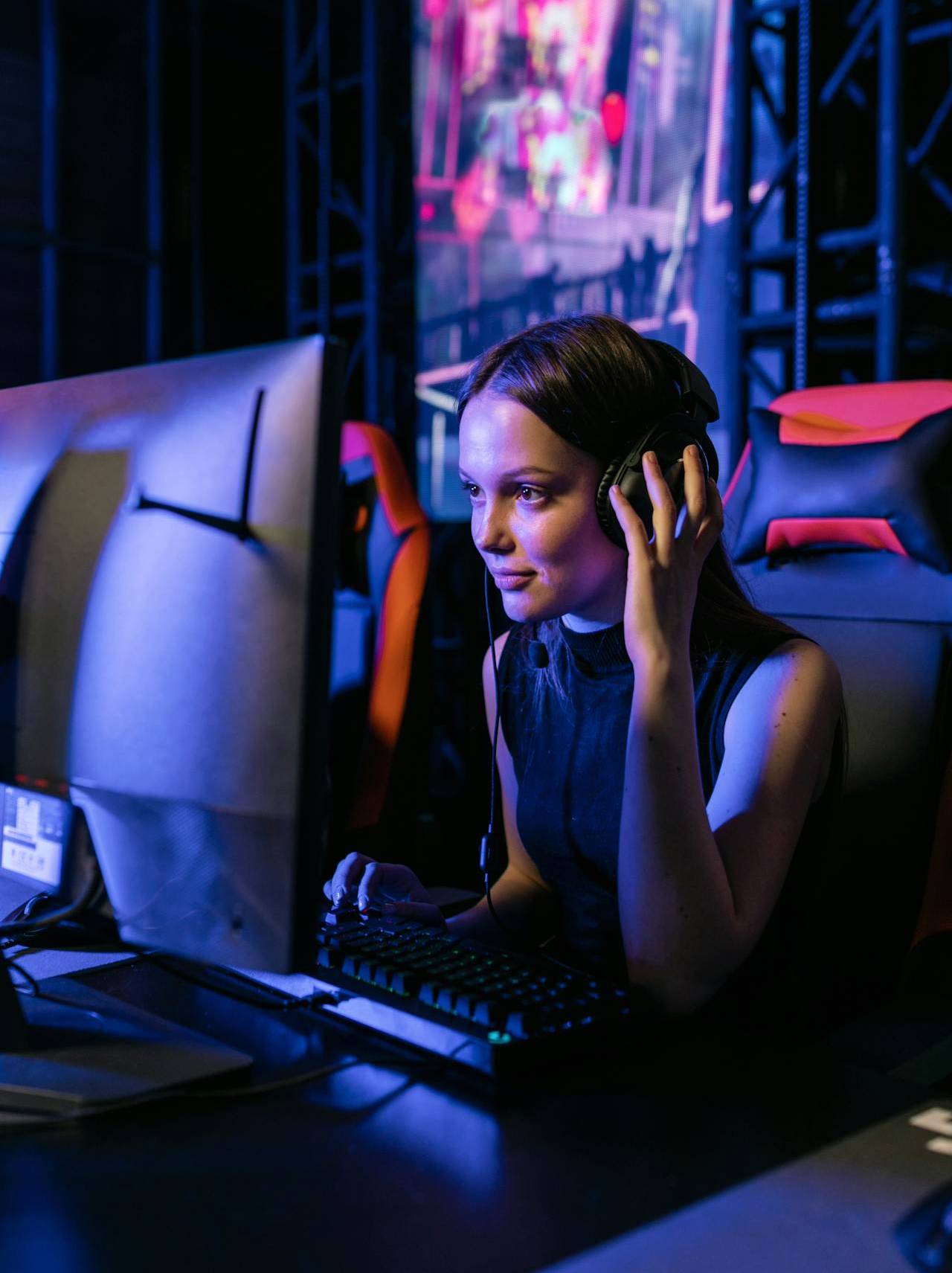 A woman with headphones is focused on her computer screen, using a video game emulator in a room with colorful lighting, indicative of a gaming or digital entertainment environment.